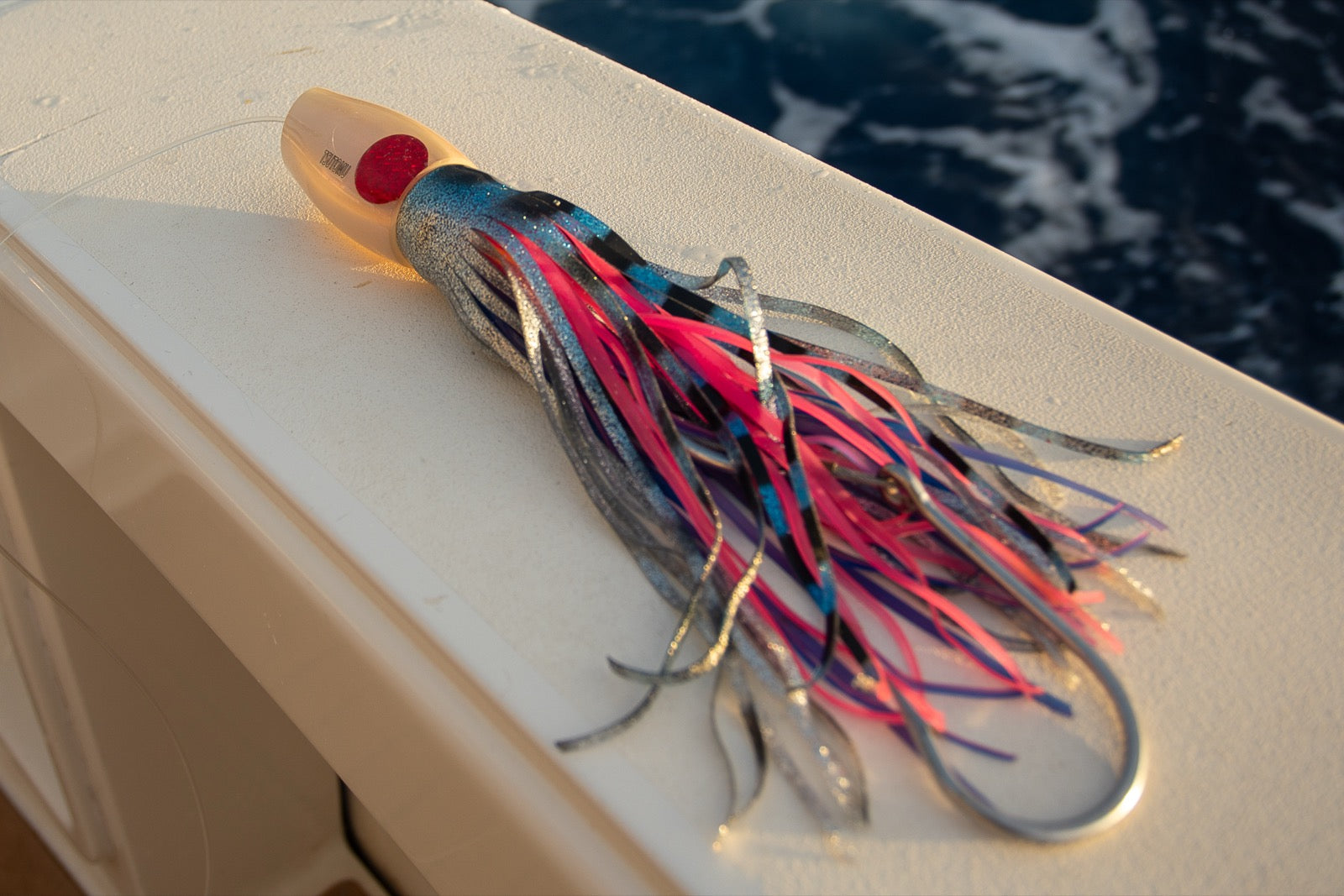 Tsutomu trolling lure with a clear bullet head and a blue, pink, and silver skirt laid out on the covering board with the water alongside the boat
