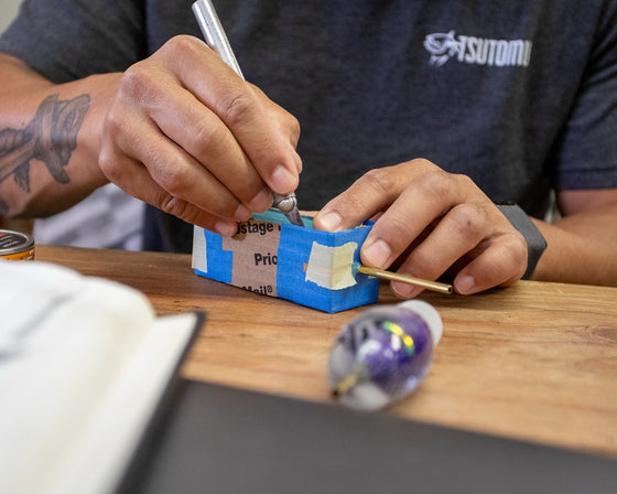 Hands at the Tsutomu shop bench working on a taped lure mold, with a finished purple lure head in the foreground